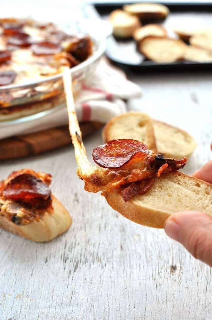 Toasted bread scooping up ultra cheesy, gooey Pizza Dip in a glass dish with a great cheese pull shot.