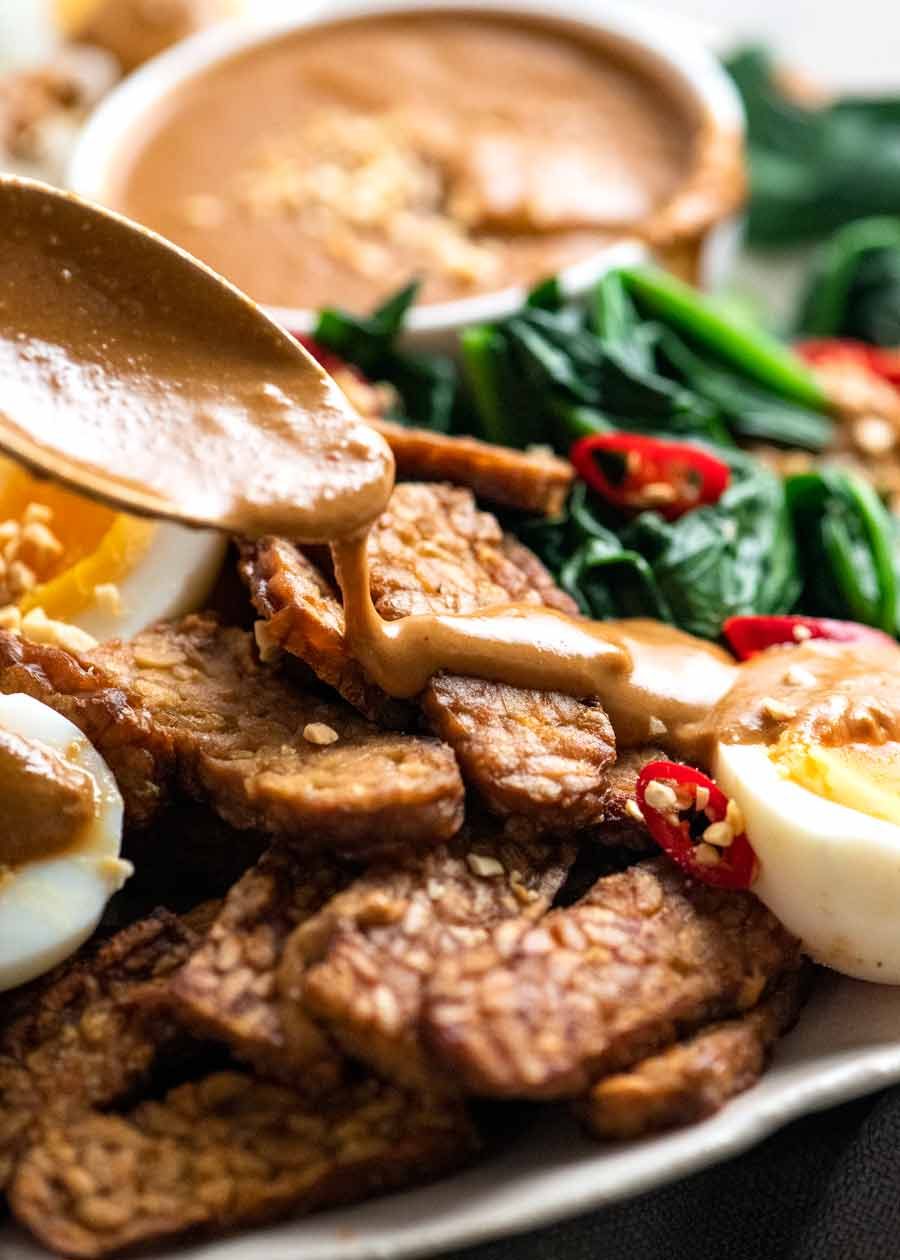 Spoon drizzling Gado Gado peanut sauce over tempeh